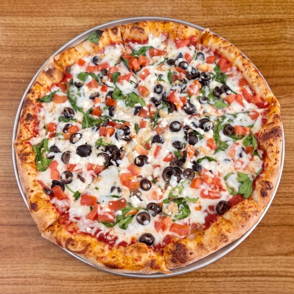 A whole pizza with white sauce, tomatoes, black olives, and spinach on a wooden table.