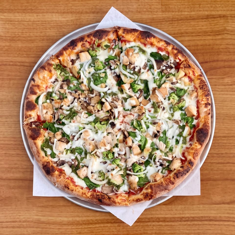 Overhead view of a whole pizza with chicken, broccoli, mushrooms, onions, and white sauce.