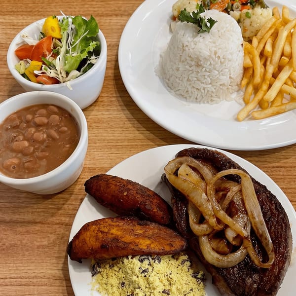 Two plates of Brazilian food, with steak, onions, plantains, rice, fries, beans, and salad.