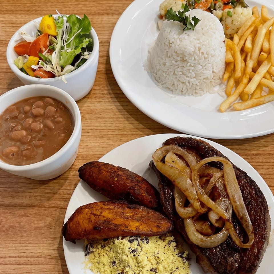 Two plates of Brazilian food, with steak, onions, plantains, rice, fries, beans, and salad.